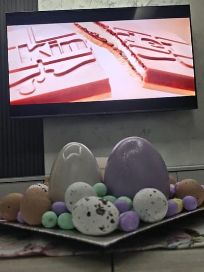 An indoor scene in Opaka, Bulgaria, captured from a slightly elevated perspective. A black-framed television screen dominates the upper portion, displaying a close-up image of two elongated, light-colored confectionery bars. The bar on the left is intact, exhibiting a light top layer with a red-brown base. The bar on the right is partially sectioned, revealing a white creamy filling between the light top and red-brown base. Embossed or molded patterns, including what appears to be the letters "KIM," are visible on the top surface of the bars. Below the television, a decorative square or rectangular tray rests on a surface covered by a floral-patterned fabric. The tray holds an assortment of decorative objects: two large, glossy egg-shaped items (one light beige, one light purple), several smaller, solid light brown egg-shaped items, and multiple white egg-shaped items speckled with dark spots. Numerous small, spherical pastel-colored objects (light green and light purple) are interspersed among the eggs on the tray. The wall behind the tray and beneath the television is a light, textured surface.