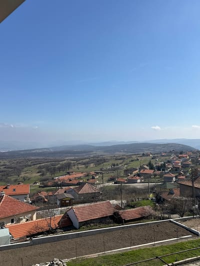 This image captures an elevated, wide view of the village of Chelopek, Bulgaria, under a clear, light blue sky with minimal, wispy clouds near the horizon. In the immediate foreground, a dark brown, tilled garden bed is visible, contained by concrete retaining walls. Green grass lines the area below and to the right of this bed. The midground features numerous houses with reddish-brown tiled roofs, typical of rural Bulgarian architecture, spread across undulating terrain. Bare deciduous trees, indicating late winter or early spring, are interspersed among the dwellings and in the surrounding fields, which show patches of green grass. Utility poles and wires are observable throughout the village. A paved road or dirt track winds through the residential areas. In the distant background, hazy rolling hills and mountains extend to the horizon. To the far left, beyond the main village cluster, faint structures suggest a more densely populated area or another town. No people are discernible, and no specific actions or interactions are visible within the frame.