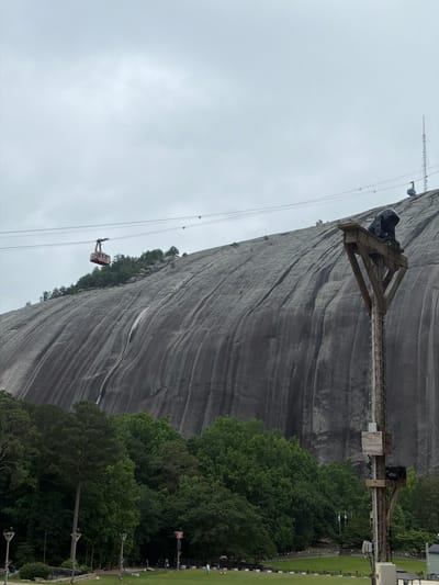 The image depicts Stone Mountain in Stone Mountain, Georgia, United States. A cable car ascends the side of the mountain. The mountain's rock face is visible. A cable support tower is positioned on the side of the mountain. Trees line the base of the mountain. The sky is overcast. An antenna is located on the peak. A patch of green grass is visible at the base of the mountain, along with utility poles.
