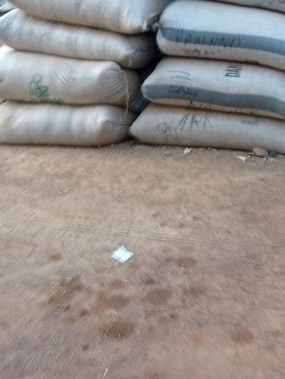The scene is an outdoor area in the city of Nassarawa, Nigeria. The upper portion of the image displays two distinct stacks of large, light-colored sacks. The stack on the left consists of four sacks piled vertically, with faint green markings visible on one sack. The stack on the right also comprises four sacks, exhibiting markings such as "JAALONG", "SAD", and additional handwritten text. These sacks appear to contain granular material. The lower two-thirds of the image depict an uneven, light brown or tan ground surface, likely compacted earth or dirt, showing various darker brown stains or damp patches. Indistinct tire or tread marks are present on the ground. A single, small, crumpled piece of white material, possibly paper or plastic, rests on the ground in the foreground. No individuals, actions, or specific interactions are visible in the frame. The lighting suggests ambient daylight.