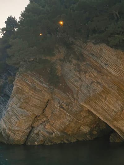 The image depicts a rocky cliff face covered in dense vegetation, with two streetlights illuminating the scene from within the foliage. The cliff exhibits distinct horizontal layering, indicating a geological formation. At the base of the cliff, dark water, likely the sea, is visible. The overall impression is of a natural coastal landscape, possibly within the vicinity of Medinski Krš, Montenegro, given the context. There are no people or explicit actions depicted in the image.