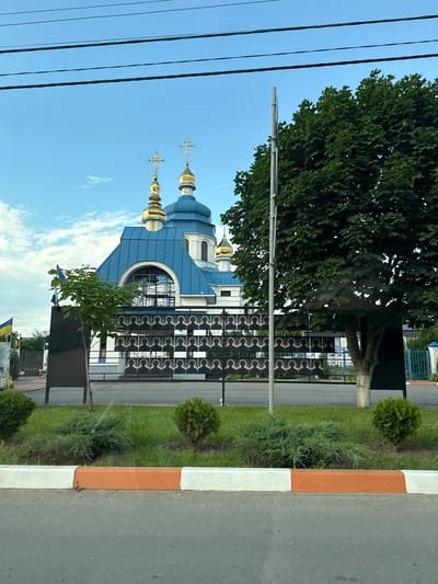 The image shows a bright, clear day with a large tree and a blue-domed church with golden crosses. The church's distinctive blue roof and golden domes suggest an Orthodox Christian place of worship. A tall fence, adorned with a repetitive decorative pattern, separates the church grounds from a sidewalk and a street. The fence is black and appears to be made of metal. The sidewalk has a curb painted with alternating orange and white stripes, a common marking in some regions to indicate pedestrian crossings or hazardous areas.
The background reveals more of the church's architecture, including windows and another dome. There's a glimpse of a smaller tree in front of the fence. Power lines stretch across the upper left portion of the image, suggesting the proximity of residential or commercial areas. The presence of a Ukrainian flag, partially visible in the lower right, places the scene in Ukraine. The overall impression is of a peaceful, well-maintained religious site on a sunny day. No people are visible in the image, and no text is legible. The time of day appears to be late afternoon or early evening, given the angle of the sunlight.