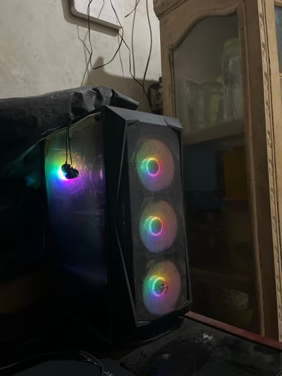A black computer tower with three illuminated fans displaying rainbow colors is sitting on a dark surface. The computer is in a dimly lit room. There is a white cabinet with glass doors in the background. The computer is positioned near the right side of the frame. The room is in a house in Kecamatan Waru, Indonesia. The location is determined from the provided coordinates. There are no people in the image.