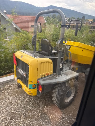 The image shows a yellow and gray dumper truck parked on a gravel surface in what appears to be a rural or semi-rural setting. In the background, there are houses with red tiled roofs, lush green trees, and a large, tree-covered mountain. The sky is overcast. The dumper truck has a visible license plate that reads "BE-818249". There is also a logo that says "De Luca" on the side of the truck. The truck is facing away from the viewer and is partially obscured by a dark, reflective surface on the right side of the image, which may be a window. The scene suggests a construction or landscaping site in or near Reconvilier, Switzerland.