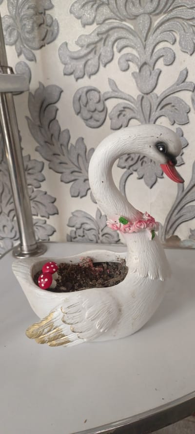 A white ceramic swan planter sits on a white surface against a backdrop of grey damask wallpaper. The swan has a pink and green floral collar around its neck and gold accents on its wings. Inside the planter, soil is visible, along with two small red and white mushroom figurines and some pink decorative elements. A portion of a metal stand is visible to the left of the swan. The image is taken in Baghdad, Uzbekistan.