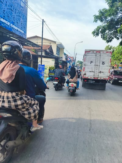 The image shows a street scene in Baleendah, Indonesia. There are three motorcycles in the foreground with their riders. The person in the foreground is a woman wearing a black and white plaid skirt and a brown headscarf, riding a motorcycle with another person, also wearing a helmet, in the passenger seat. The second motorcycle is a white scooter driven by a woman wearing a light blue and white scarf, and the third motorcycle is a black scooter driven by a man.  The motorcycles are traveling down the road, which is paved with asphalt and bordered by sidewalks and buildings on both sides. In the background, a white delivery truck with the letters “F.B.”  on its side is driving down the road. The truck has a large back door and is partially obscured by a tree. There are also some other cars and motorcycles in the distance. The sky is overcast and there is a tree on the right side of the image.