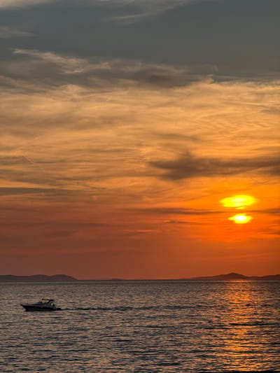 This image captures a sunset over the Adriatic Sea, with the silhouette of the Croatian coast and islands in the distance. The sky transitions from a deep blue at the top to vibrant oranges and yellows near the horizon, painted with wispy clouds. The sea below is a deep blue, reflecting the warm hues of the sunset. A small motorboat, dark against the water, is moving from left to right, leaving a faint wake. The scene is set in Primošten, Croatia, known for its picturesque coastal landscapes.
