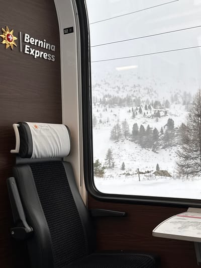 Inside the Bernina Express, a window reveals a snow-covered landscape dotted with trees. The scene outside the train is obscured by fog. Beside the window is a black seat, with a white, cylindrical container resting on the floor next to it. The train car's interior is modern and clean. The name "Bernina Express" is printed on the wall next to a Swiss flag emblem.