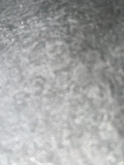 The image displays a blurred, out-of-focus texture that appears to be a metallic or reflective surface. There are numerous small, circular highlights scattered throughout, suggesting a shimmering or glittering effect. The overall color palette is predominantly gray and white, with some subtle hints of iridescent colors within the highlights. The texture seems to be uneven, with variations in light and shadow creating a sense of depth. There are no discernible objects, people, or specific environmental features visible due to the extreme blurriness of the image. The location in Ablanitsa, Bulgaria, cannot be determined from the visual information provided in the image.