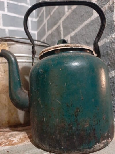A close-up, eye-level shot shows a weathered, dark teal kettle with a black metal handle. The kettle is positioned in the foreground, slightly off-center to the right. Its surface is marked with rust and some lighter scuffs, suggesting age and use. The spout of the kettle extends to the left. Behind it, a portion of a lighter colored metal container is visible. The background consists of a grey brick-patterned wall. The lighting is somewhat dim, with a reflection visible on the kettle's surface. The overall impression is one of a utilitarian, perhaps vintage, kitchen or storage area. There is no indication of people or specific actions taking place. The scene is indoors, in Zalesovo, Russia.