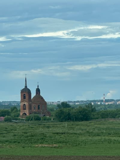 The image, taken from a low angle in Татарские Челны, Russia, depicts an old, brick church with two prominent domes, one pointed and the other rounded. The church appears to be in a state of disrepair, with some visible crumbling and moss growth. It is set against a backdrop of a vast, clear blue sky with streaky white clouds. To the right of the church, a green, grassy field stretches into the distance, with a line of trees forming a border further back. In the far distance, a cityscape is visible, including a tall, red and white striped chimney, suggesting an industrial presence. The overall impression is one of a rural, slightly weathered, historical site under a bright, possibly late afternoon, sky. No people or specific activities are visible, and there is no visible text.