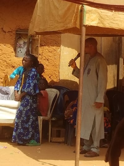 This image captures an outdoor scene in Ochadam, Nigeria, depicting a gathering with several individuals under a temporary fabric canopy.
In the center-right, a man stands, partially obscured by a tent pole, facing left and holding a black microphone to his mouth. He is dressed in a light grey long-sleeved tunic (kaftan) with a dark embroidered pattern on the left chest, matching trousers, and dark brown sandals.
To the left, a young girl stands prominently, facing forward and slightly left, looking upward. She wears a long blue dress with a geometric pattern and bright green sneakers. Behind her, another young child, wearing a red shirt, is partially visible, looking towards the right. Another child in blue clothing is partially visible behind the girl in blue.
The background consists of a rough, orange-brown mud-plastered building wall with a small, open window containing an unclear image or object. On the far left, a light-colored upholstered bench or bed frame is visible with white fabric. Behind the children, there are white and dark blue plastic monobloc chairs.
The ground is reddish-brown earth, indicating an outdoor setting, with some small debris present. A light-colored fabric canopy is stretched overhead, supported by dark poles. The overall impression is a community gathering or event taking place during daylight hours.