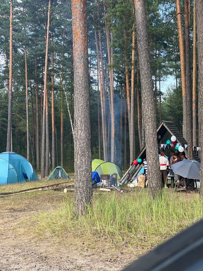 A group of people are gathered at a campsite in a pine forest. Several tents are set up, including a large blue tent to the left, a smaller green tent in the middle, and a green and yellow tent to the right. A rustic wooden structure with a pitched roof stands in the center, adorned with balloons and bunting. Several individuals are interacting around this structure, with some seated under a grey umbrella to the right. Smoke is visible rising from a fire in the middle ground, indicating a campfire. The ground is covered with sand, dry grass, and pine needles. The overall setting suggests a recreational gathering or event in a natural environment within the Дражнаўскі сельскі Савет, Belarus.