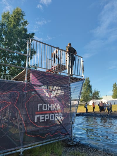 An outdoor scene features a temporary structure, likely part of an obstacle course, positioned adjacent to a body of water. The structure is constructed from metal scaffolding and includes a platform with a railing, from which two individuals are visible. One person, dressed in camouflage attire, stands facing away from the camera, leaning on the railing. Another individual, wearing a white shirt and dark pants, is partially visible on the platform, descending a set of stairs.
A large banner is draped across the front of the structure, displaying the Russian text "ЛИГА ГЕРОЕВ" and "ГОНКА ГЕРОЕВ," which translates to "League of Heroes" and "Race of Heroes." The banner has a dark background with red topographic-like patterns.
In the background, several people are gathered near white tents, suggesting an event or competition is taking place. The environment is grassy with scattered trees, and the sky is clear blue with scattered clouds.
The scene is located at the Sertolovo Sports Complex in Sertolovo, Russia, a venue known for hosting the "Race of Heroes" obstacle course events.