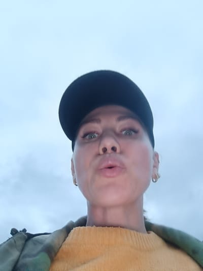 A low-angle, medium shot captures a woman wearing a black baseball cap and a yellow sweater. Her lips are pursed, and she is looking forward. Her left ear is visible and adorned with a small hoop earring. She is wearing a camouflage jacket over her sweater. The background is a cloudy sky, suggesting an outdoor setting. The image does not contain any information to determine its location in Verkhnie Chershily, Russia.