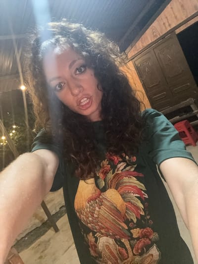 A selfie photograph, likely taken outdoors at night in what is perhaps the city of Bắc Hà, Vietnam, based on the prompt. The image features a person with curly dark hair, light skin, and visible teeth, looking down at the camera. The person is wearing a dark-colored t-shirt with a floral or avian design. In the background, there are dim lights, possibly string lights, and an out-of-focus building or structure that appears to be made of wood or metal. A red crate-like object is also faintly visible in the upper portion of the background. The ground is a pale, possibly concrete or stone, surface. The overall impression is one of low-light conditions and an informal setting.
