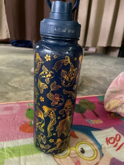 A close-up, slightly high-angle shot shows a dark blue water bottle adorned with golden koi fish and flower designs. The bottle is lying horizontally on a surface that appears to be a children's playmat with cartoon figures and bright colors. The brand name "AquaFlask" is visible on the blue cap of the bottle. The background includes a glimpse of light-colored wooden slats and a partially visible object covered in pink fabric. The image was taken indoors. Hijo, Philippines cannot be determined from this single image.