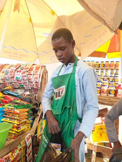 In the city of Kawaji, Nigeria, a young male individual is positioned at an outdoor market stall, primarily engaged with items at waist height. He wears a light blue long-sleeved shirt and a green apron featuring the "Nestle MILO" logo. His head is slightly bowed, and his gaze is directed downwards as he uses both hands to hold a black plastic bag, which contains smaller packaged goods visible inside.
Above the scene, a large yellow market umbrella or awning provides shade, displaying inverted text, including phrases like "Enjoy the taste." To the left of the individual, numerous small, colorful sachets and packaged items are stacked on a counter and displayed vertically on a wooden frame. A green plastic bowl is partially visible in the lower left. To the right, behind the individual, multiple rows of jars and bottles, likely containing condiments such as mayonnaise and sauces, are arranged on wooden shelves. The lower body of another individual, wearing light-colored pants and appearing barefoot, is partially visible in the far right foreground.
