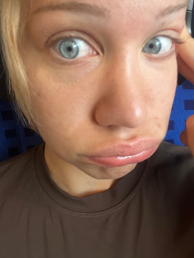 A close-up, front-facing photograph captures a young Caucasian woman with blonde hair and striking blue eyes. She is wearing a dark brown t-shirt and her face is turned slightly to the left, with her lips pursed outwards. Her right hand is visible touching her right cheek and hair. The background consists of a blurry blue surface with a dark checkered pattern. The lighting suggests an indoor setting. The image does not contain any specific visual cues to indicate a location within the city of Ampfing, Germany.