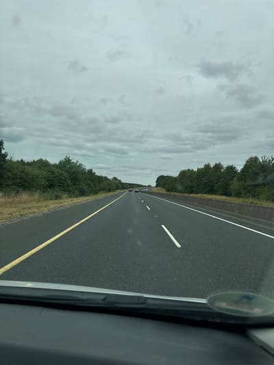 The image depicts a point-of-view shot from inside a vehicle on a multi-lane highway. The road is asphalt with clear lane markings, including a solid yellow line on the left and dashed white lines separating lanes. Trees and vegetation line both sides of the highway, with a concrete barrier visible on the right. The sky is overcast with heavy, grey clouds. Several vehicles are visible on the highway in the distance, traveling in the same direction. The scene appears to be a stretch of highway in the rural area surrounding Ballyduff South, Ireland.