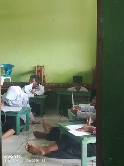 The image depicts an interior scene, identified as a classroom in Wanguk, Indonesia. Five individuals, presumed students, are present. Multiple individuals are seated on the white tiled floor at small, dark green wooden desks. In the foreground, two sets of bare feet are visible on the floor; one person's hands are actively writing on a white paper with a pen at a desk. A student wearing a white head covering leans forward over their desk. In the middle-ground, a student in a white shirt holds a paper near their mouth, while another student, wearing a black peci, sits upright behind a desk, looking forward. The classroom features light green painted walls with visible marks and partial, inverted text. A blue and white plastic chair and stacked wooden frames are positioned against the back wall. A dark vertical wooden frame partially obstructs the right side of the view.