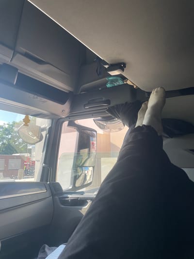 The image, taken from inside a truck cab, shows a person's legs and feet, clad in white socks and dark pants, propped up on the dashboard. The sun is shining through the windshield, casting light on the interior. Outside the truck, a building with pink walls and windows is visible, along with trees and other structures. A yellow tree-shaped air freshener hangs from the rearview mirror. The scene appears to be a daytime setting in an urban or semi-urban environment.