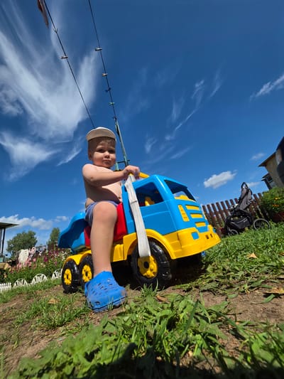 A young child wearing a white cap and blue Crocs sits on a blue and yellow toy dump truck outdoors in a grassy yard. The child is shirtless and wearing blue shorts. The truck has large black and yellow wheels. Behind the child, a brown wooden fence separates the yard from a garden with pink and purple flowers. A gray baby stroller is parked next to the fence. The sky is bright blue with wispy white clouds. Several clothespins hang on a wire in the upper left portion of the image. The scene appears to be in a rural or suburban setting in Буренка, Russia, given the style of the house and garden.