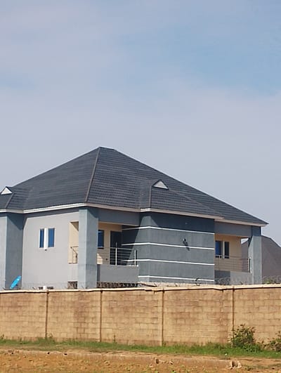 This is a photograph of a modern, multi-story house with a dark grey, sloped roof, taken from a low angle, possibly in the city of DU, Nigeria. The house has a light grey and dark grey exterior with horizontal white trim. There are several windows visible, some with blue tinting. A beige-colored wall with a barbed wire fence runs across the foreground, separating the property from the viewer's perspective. The ground in front of the wall appears to be dry, dusty earth with some sparse vegetation. The sky is a clear, pale blue. There are no people or animals visible in the image.
