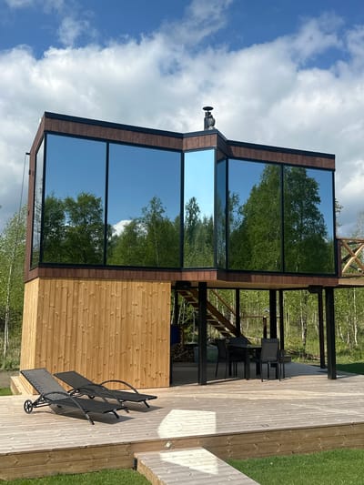 The image shows a modern building located outdoors in a wooded area near Vaibla, Estonia. The building is constructed with a wooden exterior, accented by large, vertically aligned mirrored windows reflecting the surrounding trees and sky. A chimney is visible on the roof. A wooden deck extends from the building, furnished with dark-colored lounge chairs and an outdoor dining set under a metal frame structure. The deck transitions to a grassy lawn. The photograph is taken in daylight, with some clouds in the sky.