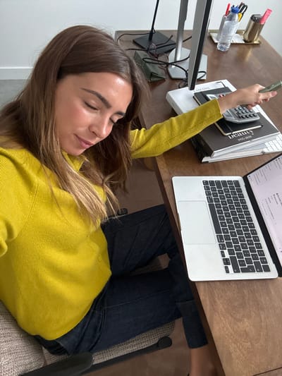 A woman in a yellow sweater is lying down at a desk. She is holding a small, grey object in her hand. On the desk is a laptop, a stack of books, a calculator, and a water bottle. The woman is wearing dark jeans and sitting in a grey chair. There is no indication that the image was taken in Torres Vedras, Portugal.
