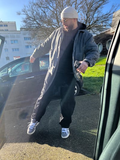 An adult male is depicted outdoors in Hérouville-Saint-Clair, France, on a clear, sunny day. The subject, with a beard, wears a light-colored beanie, round sunglasses, a dark grey jacket over a black hooded sweatshirt and t-shirt, black pants, and grey and white sneakers. He is captured mid-motion, facing towards the right foreground, with his mouth slightly open. He holds a dark-colored smartphone with three camera lenses in his left hand. To the left, a dark compact car is parked on an asphalt surface, its driver-side door partially open, reflecting sunlight. A floral-patterned sticker is visible on the car's rear passenger window. In the background, a multi-story, light-colored residential building is present to the left, while a wooden building with two circular windows and green grass is visible to the right. Bare deciduous trees stand against a clear blue sky.