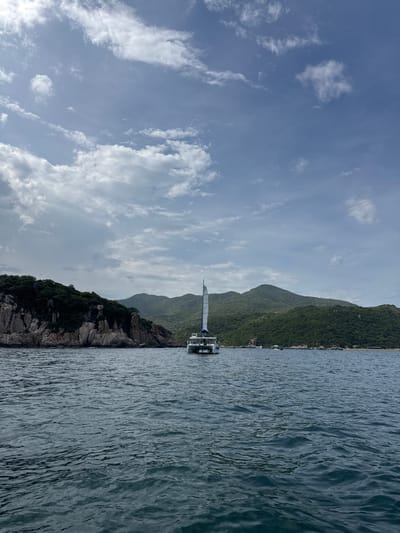 An image captures a catamaran sailing on dark blue water, with lush green hills in the background and a rocky coastline on the left. The sky above is a mixture of blue and white clouds. The catamaran, with a white hull and sail, is positioned in the center of the frame, moving away from the viewer. The rocky island to the left is densely covered with trees and vegetation. The hills in the background are also covered in dense green foliage. The water is slightly choppy, indicating moderate wind conditions. The overall scene depicts a serene coastal landscape with a boat on the water. This scene is characteristic of the coastal areas near Vinh Hai, Vietnam, known for their natural beauty and water activities.