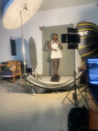 A young woman stands on a pedestal in a studio setting, posing for a photograph. She is wearing a white dress with navy blue polka dots and dark open-toed heels. Her hands are on her hips, and she looks towards the camera with a slight smile. The studio is equipped with professional photography equipment, including a large softbox with a black and yellow grid on the left, a ring light in front of the woman, and a backdrop. A monitor displaying an image is visible in the background, along with a black bag and a wooden chair. The floor is tiled. The lighting suggests a controlled studio environment, likely indoors during the daytime. The woman's posture and expression convey confidence and readiness for the photoshoot. The overall scene depicts a professional photography session in progress.