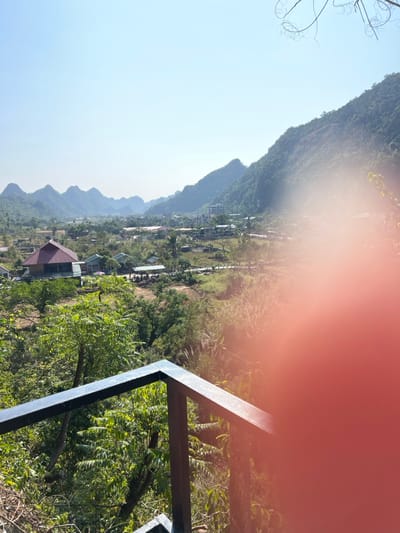 The image displays a wide landscape view of Trân Châu, Vietnam, centered on a valley or basin framed by majestic, tree-covered karstic mountains. The sky is clear and a bright, light blue, indicating a sunny day. In the lower right foreground, a dark metal railing or frame is partially visible, suggesting the photo was taken from an elevated vantage point, such as a balcony or lookout platform. Sparse, bare tree branches are also seen in the upper left corner.
The mid-ground reveals a sprawling rural settlement, comprising numerous houses and buildings nestled amidst abundant green vegetation. Many structures feature distinct reddish-brown roofs, while others have lighter or metallic coverings. Lush trees and dense foliage cover the slopes of the mountains and are scattered throughout the valley, contributing to a vibrant natural setting. The distant mountains recede into a soft atmospheric haze.
A significant reddish-orange lens flare partially obscures the upper right quadrant of the image, likely caused by direct sunlight entering the lens. The scene conveys a sense of natural beauty and tranquility. No people are visible, and there is no discernible text within the frame.