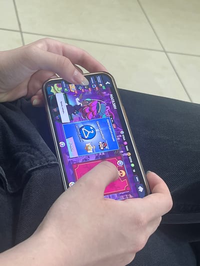 A close-up, eye-level shot captures a person holding a smartphone displaying a video game. The phone, with a gold-colored frame, is held in landscape orientation. The game interface features vibrant purple and pink colors, with cartoonish characters and text in Russian. A prominent text on the screen reads "МАГАЗИН" (Shop). The person's hands, with neatly manicured nails, are positioned to interact with the phone. The left hand is slightly obscuring the bottom left corner of the screen, while the right thumb is pressing on the screen. The background consists of a tiled floor with light-colored square tiles and dark grout lines. The image is taken indoors, likely in a public space or home. The context of Boksitogorsk, Russia, is not evident from the visual information presented in the image itself.