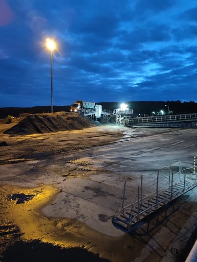 The image depicts a nighttime scene at an industrial site, likely a construction or material processing yard, under a dramatically cloudy, deep blue sky. A tall lamppost with a bright, glowing light dominates the left side of the frame, casting a warm illumination on the surroundings. In the middle ground, a large pile of dark material, possibly dirt or gravel, is visible. A yellow truck is positioned on an elevated conveyor system, which appears to be in operation, with lights illuminating the equipment. The ground in the foreground is a mix of concrete and wet, reflective surfaces, with tire tracks indicating vehicle activity. A metal walkway with handrails and rope barriers is prominently featured in the lower right quadrant, casting long shadows. The background shows a dark, tree-lined horizon with scattered lights, suggesting the presence of a settlement or further industrial activity. The overall atmosphere is one of ongoing work under artificial light in a natural environment at dusk or dawn.
