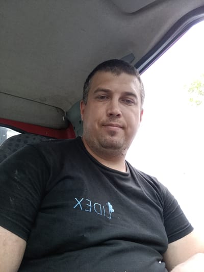 A man with short brown hair and a light beard is photographed from the chest up, sitting inside what appears to be a vehicle. He is wearing a black t-shirt with a light blue logo consisting of the letters "IDEX" and a stylized butterfly or flower. The man is looking directly at the camera with a neutral expression. The interior of the vehicle is visible, including the grey headliner and a red door panel. The background outside the vehicle is a bright, overexposed sky with hints of green foliage. The photograph was taken in Svémyslice, Czechia.