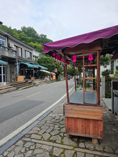 The image captures a street scene in Xihu District, China. In the foreground, a wooden market stall with a purple awning stands on a cobblestone sidewalk. The stall is empty, with a grey mat on its floor and several pink decorative items hanging from its roof. To the left of the stall is a paved road with white and yellow lane markings. Along the road, there are buildings with awnings and shops. In the background, a lush green hillside is visible, with trees covering its slope. The sky is overcast. There are no people clearly visible in the immediate foreground, but figures can be seen further down the street, and a scooter is parked near the stall. The overall atmosphere is that of a quiet street in a town or village.