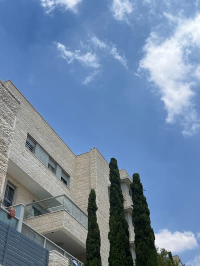 This is a low-angle, eye-level shot of the exterior of a modern building in Modiin-Maccabim-Reut, Israel. The building is constructed with light-colored stone blocks and features multiple balconies with glass railings. Tall, dark green cypress trees stand in front of the building, partially obscuring the lower floors. The sky is a bright blue with scattered white clouds. The scene appears to be captured during daylight hours under clear weather conditions. There are no people or visible actions depicted in the image.