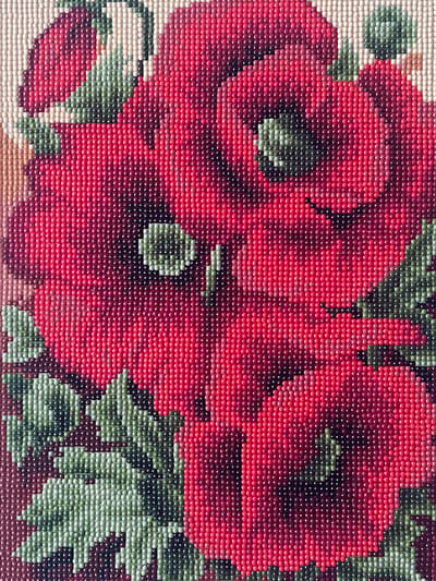 The image displays a close-up view of a cross-stitch or diamond painting depicting a cluster of red poppies with green leaves. The artwork is rendered in small, uniformly colored dots, creating a pixelated effect. The poppies are in various stages of bloom, with visible stamen and petals. The lighting suggests a bright, even illumination, highlighting the texture of the individual dots. There are no people, animals, or identifiable landmarks present in the image. Therefore, its location in Sherbakul, Russia, cannot be determined from the visual information provided.