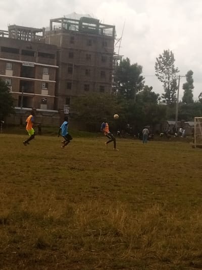 The image depicts an outdoor football (soccer) scene in Karingani ward, Kenya, under an overcast sky. Three male individuals are actively playing football on a field with patchy, brownish-green grass in the foreground. The leftmost player wears an orange vest over a light green shirt and dark pants, with his left leg raised. The middle player wears a light blue shirt and dark pants, positioned with bent knees. The rightmost player, in an orange shirt and dark pants, is in the process of kicking or controlling a light-colored football, which is suspended in mid-air near his right foot.
In the background, on the left, a multi-story, brownish-grey building with multiple windows is visible, bearing faint text on its side. Behind and slightly to the right of this structure, a taller, multi-story building is under construction, displaying exposed concrete and scaffolding elements on its upper levels. A dense line of green trees and bushes extends across the middle ground, separating the field from the background structures. To the right background, a group of approximately 10-15 individuals are gathered near additional trees and a white-framed football goalpost. A utility pole with wires is also visible on the far right edge of the frame.