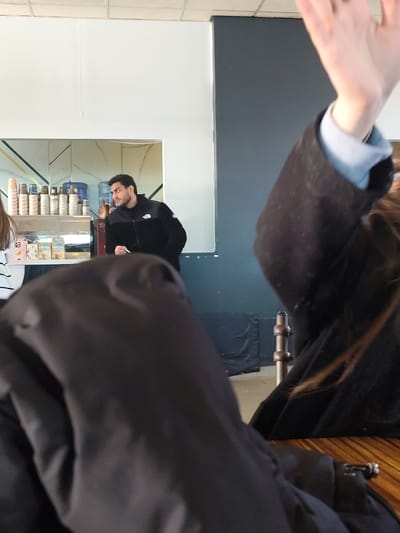 A cafe scene shows a man in a black jacket standing behind a counter stocked with coffee cups and condiments. He looks to his left. In the foreground, a person with their hand raised is partially blocking the view. Another person, obscured by a large black jacket, is seated at a wooden table. Background details include a gray wall and a curtained area.