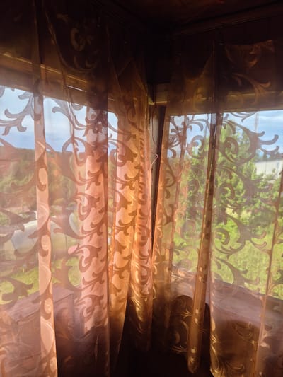 This media file depicts a close-up view of sheer, patterned curtains, likely made of a translucent, flowing fabric. The curtains are a warm, coppery-brown hue and are adorned with an ornate, swirling damask pattern that is darker than the base fabric. Sunlight streams through the curtains, creating highlights and shadows and revealing the pattern in varying degrees of opacity.
Through the translucent fabric, a bright, outdoor scene is visible. Hints of blue sky suggest it is daytime, and the presence of lush green foliage, appearing as a soft blur, indicates a location with trees and vegetation. The light filtering through suggests it might be a sunny day.
The curtains are arranged in multiple horizontal folds, creating a layered effect. The way the fabric drapes suggests it is hung in a window, possibly in a residential setting. The wooden structure framing the window is partially visible on the left and right edges of the image, adding to the sense of being indoors looking out. A thin, pink wire or string is visible across the bottom portion of the image, caught on the curtain or the window frame, adding a small, unexpected detail.
The overall mood is serene and slightly dreamy, with the soft light and flowing fabric creating a gentle, diffused atmosphere. The detailed pattern on the curtains adds a touch of elegance. There is no visible text in the image. The setting appears to be a domestic interior in Mashzezero, Russia, looking out onto a verdant outdoor environment.