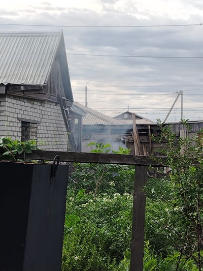 The image depicts a cloudy outdoor scene, likely in a rural or suburban area of Ekibastuz, Kazakhstan. In the foreground, a dark metal object, possibly a bin or container, is visible on the left. Behind it, a wire mesh fence runs horizontally across the frame, separating it from a dense area of green vegetation with scattered white flowers. Several wooden posts support the fence.
In the midground, there are elements of residential structures. A white brick building with a dark opening is on the left, partially obscured by foliage. To the right of this, a wooden structure with a sloped roof and visible beams is present, with smoke emanating from behind it. A ladder leans against this structure. Further to the right, another building with a corrugated metal roof is visible, along with utility poles and overhead wires that traverse the sky. The overall environment suggests a yard or garden area adjacent to buildings, with evidence of potential activity such as a fire or cooking, indicated by the smoke. The sky is overcast, with layers of grey clouds.