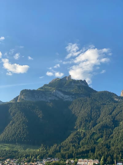 An eye-level, wide shot captures a densely forested mountain range under a clear blue sky with scattered white clouds. The mountain's peak is rocky, with a sharp, spire-like formation. A village with white and light-colored buildings is nestled at the base of the mountain, partially obscured by trees. The terrain is steep and covered in a thick canopy of dark green coniferous trees, with some areas of lighter green indicating cleared patches or different vegetation. The sun is casting shadows on the mountain, suggesting it is late afternoon or early morning.