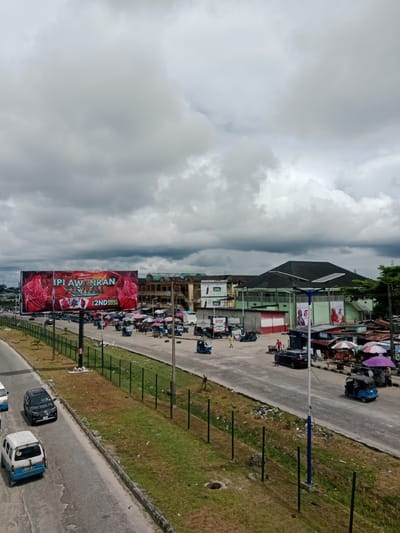 This image depicts an outdoor scene in Ekurede-itsekiri, Nigeria. The sky is filled with heavy, dark clouds. In the foreground, a paved road is visible, with several vehicles including a black car, a white and blue van, and a few blue three-wheeled auto-rickshaws, also known as "keke napep." These vehicles are either parked or in motion. A green fence runs along the side of the road, separating it from a grassy area littered with debris.
A prominent billboard with red and white graphics advertises the "IPI AWANRAN FESTIVAL" and mentions "2ND AUGUST 2026." Further down the road, a marketplace is set up with numerous stalls covered by colorful umbrellas. Several people are seen browsing or operating these stalls. In the background, various buildings are visible, including a large green building with a dark roof and a billboard advertising a "GREEN ACTION" product. The overall atmosphere suggests a bustling urban environment on a cloudy day.