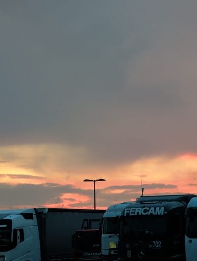 A low-angle, full shot shows a row of parked semi-trucks against a dramatic sky at dusk in Denkendorf, Germany. The sky is filled with streaks of orange, pink, and gray clouds, with a lamppost visible in the upper right quadrant. The trucks are dark and silhouetted, with some logos and numbers visible. The Fercam logo is prominent on one of the trucks, along with the number 702. Another truck to the left is white, and the truck to the far right is partially visible. The scene suggests a transportation hub or a truck stop.