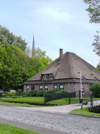 The image presents a house with a thatched roof, likely situated in Oudorp, Netherlands. The house is constructed of brick and features multiple windows with dark shutters. A chimney rises from the roof, which is covered in dense thatch material. A flagpole stands near the house. The structure is partially obscured by trees. A church steeple can be seen in the background. A short, dark fence borders the property, and an ornate metal gate provides access. The street in front of the house is paved with cobblestones. The sky is overcast. A yellow car is parked nearby.