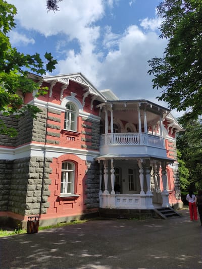 The image shows the historic building of the Central Library of Borjomi, Georgia, located on a tree-lined street. The building is constructed with a combination of pink brick and grey stone, featuring ornate white wooden railings on its porch. The year "1895" is inscribed on the facade, indicating its construction date. The sky is partly cloudy with a bright blue expanse. Green trees with dense foliage surround the building, casting shadows on the paved ground in front. Two women are visible in the foreground, one wearing red pants and a white shirt, and the other in a dark top and dark pants, standing on the steps leading to the porch. A trash bin is located to the left of the building. The overall scene depicts a sunny day in a park-like setting, highlighting the architectural beauty of the library.