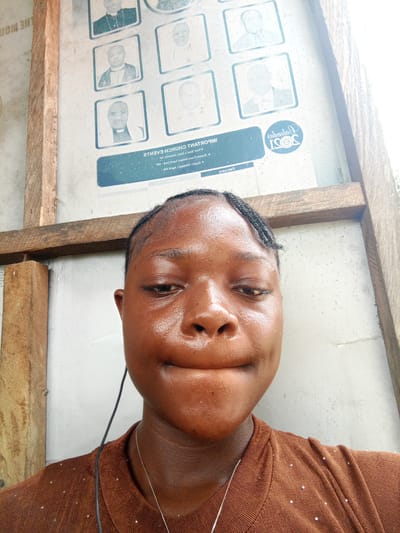 A close-up, high-angle selfie shows a young, dark-skinned woman wearing a brown top and a silver necklace. She is positioned in front of a wall featuring framed photos of clergy members and text in both forward and backward print. The text includes "IMPORTANT CHURCH EVENTS" and "JOSI", as well as information written upside down. A wooden frame surrounds the wall. The scene is in Mbribit, Nigeria.